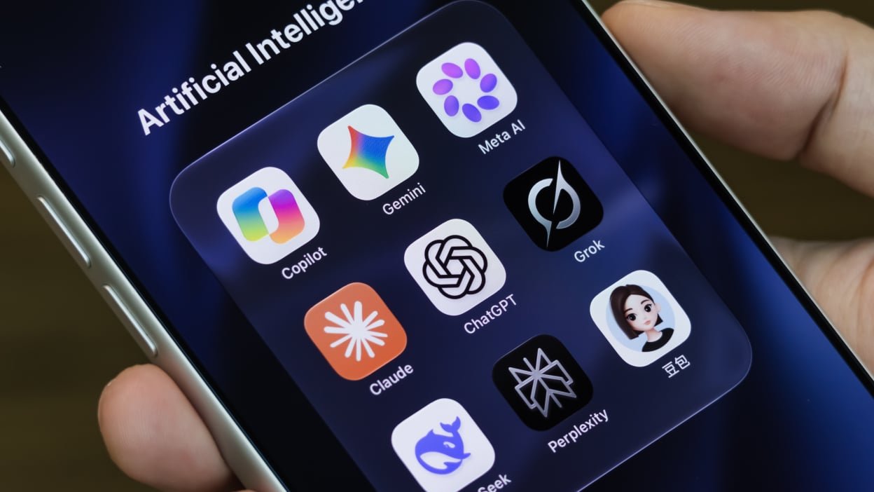 a person holds an iPhone displaying icons for Copilot, Gemini, Meta AI, Claude, ChatGPT, Grok, DeepSeek, Perplexity and Doubao in a folder titled "Artificial Intelligence"